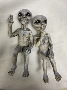 Top view of two grey resin alien figurines in sitting poses making V-sign and shushing gestures
