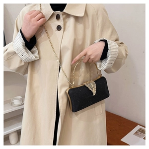 Detail of black clutch bag with golden wing clasp and chain strap worn on a trench coat