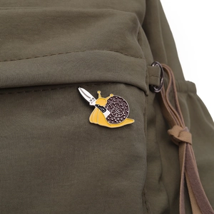 Detail view of a funny yellow snail enamel pin with a silver knife on a green backpack