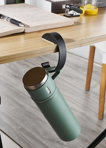 Detail of a black desk hook holding a green thermal flask on a wooden table edge