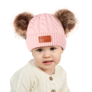 Pink knit baby beanie with two brown pom-poms and leather patch worn by an infant