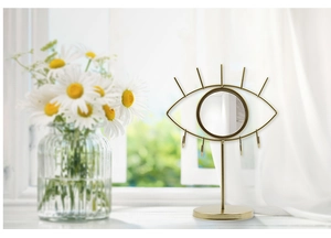 Golden eye-shaped jewelry display stand with central mirror and hooks on a bright dresser