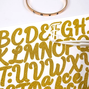 Close-up of 1-inch gold glitter alphabet stickers being applied with tweezers for DIY crafts