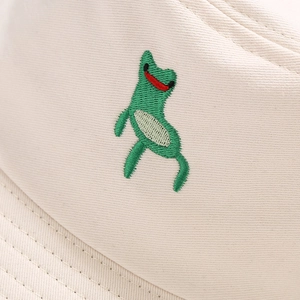 Close-up of green frog embroidery detail on beige cotton twill bucket hat texture