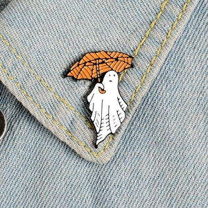 Close-up of a white ghost enamel pin holding an orange umbrella on blue denim fabric