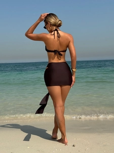 Three-piece dark brown bikini set featuring a wrap skirt shown against a beach background