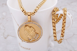Detail of 18K gold stainless steel prayer hands pendant necklace and box chain on a cup