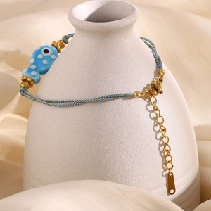 Detail of woven light blue cord bracelet with 18K gold fish charm on a white vase