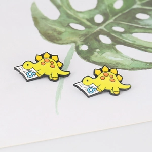 Detail of yellow stegosaurus enamel pin featuring a dinosaur reading a book on leaf decor