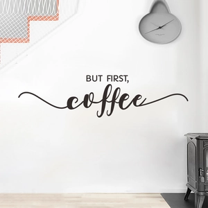 Modern black 'BUT FIRST, coffee' wall sticker on a white wall in a cozy home setting