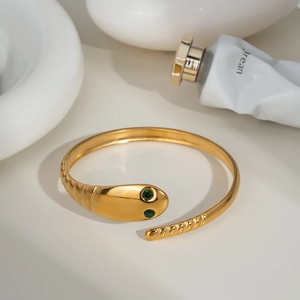 Minimalist gold snake-shaped open bracelet with green gemstone eyes on white ceramic