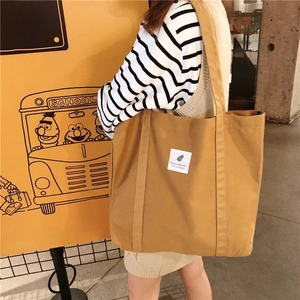 Mustard yellow canvas tote bag with logo patch shown on shoulder in a lifestyle setting