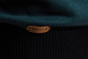 Detail von dunkelgrünem Strickstoff mit schwarzem Rippsaum und Leder-Logo-Patch