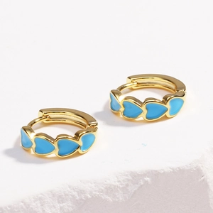 Detail shot of golden hoop earrings featuring blue enamel heart charms on a white surface