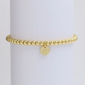 Detailed view of golden beaded bracelet featuring a round metal tag engraved with letter C