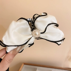 Detail view of a cream satin bow hair claw with pearl center and black woven piping