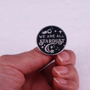 Black enamel pin featuring WE ARE ALL STARDUST text with stars and comets design