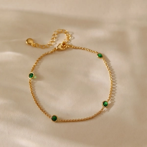 Elegante pulsera de cadena dorada con circonitas verdes esmeralda sobre fondo beige