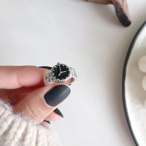 Macro detail of a silver watch-shaped ring with a black dial worn on a finger