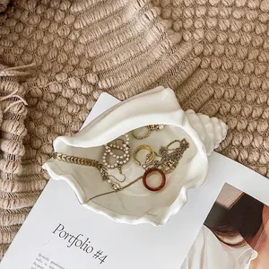 White ceramic conch shell jewelry tray on a white magazine with golden accessories