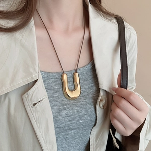Dettaglio di una collana con pendente a U in metallo dorato su modella con cappotto beige