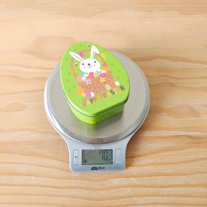 Easter egg-shaped tin box weighing 70.3g on a scale, showing bunny and egg pattern