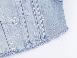 Détail d'une veste en jean bleu clair avec perles blanches, strass et ourlet effiloché