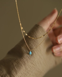 Détail d'un collier double rang doré avec pendentif goutte en opale bleue sur fond en lin