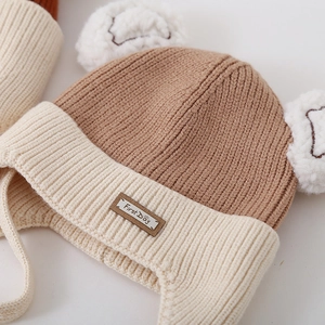 Detail of baby knit hat with ear flaps, pom-poms and First Day decorative patch