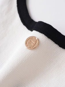 Detail eines weißen Strickoberteils mit schwarzem Rand und Metallknopf mit K-Motiv