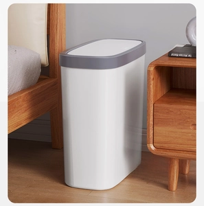Slim white trash can with grey lid placed in a narrow bedroom gap near a wooden bed frame