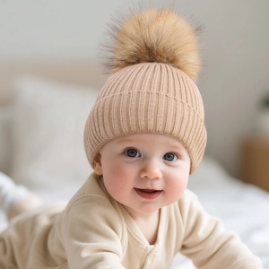 Infant beige knitted beanie with a faux fur pompom shown on a baby in a cozy setting