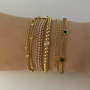 Stack of gold layered bracelets featuring beaded chains and a green emerald-cut gemstone