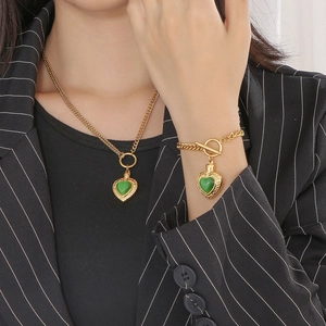 Gros plan d'une chaîne dorée avec fermoir OT et pendentifs cœur vert sur collier et bracelet