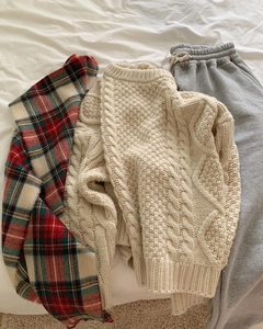 Flat lay detail of a red plaid tassel scarf with a white cable knit sweater on sheets