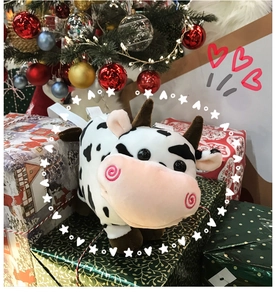 Plush cow shoulder bag on a gift box near a decorated Christmas tree background