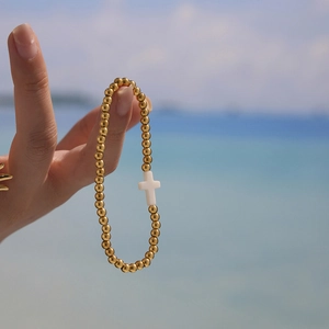 Handmade 18K gold-plated beaded bracelet with white cross charm worn on beach background