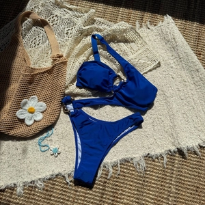 Flat lay of navy blue bikini with metal ring detail on white tassel rug for beach vacation
