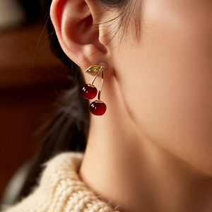 Macro shot of red cherry drop earrings with gold stems and green leaf zircons on ear