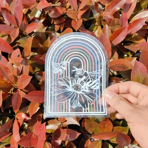 Detailed suncatcher sticker with rainbow, bee and flower design held against green leaves