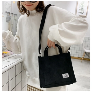 Close-up of black corduroy tote bag with white woven tag worn by a model in a bright room