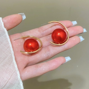 Elegant gold C-shaped hoop earrings featuring large red pearls shown in a hand-held view