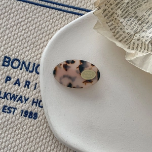 Close-up of tortoiseshell acetate hair clip with golden brand logo on a textured surface