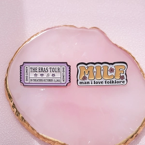 Colorful enamel pins featuring Eras Tour ticket and MILF design on pink tray
