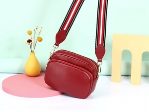 Detail van rode crossbodytas van generfd leer met gestreepte riem en drie ritsen