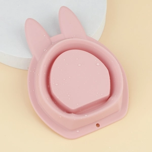 Pink rabbit-shaped silicone cleaning bowl with water droplets on a geometric stone background