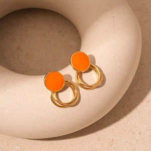 Detail of geometric earrings with orange enamel disc and triple gold rings on beige props
