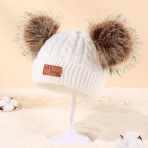 Detail of white cable knit baby hat with double faux fur pom-poms and leather patch