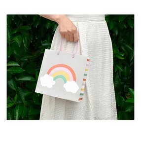 Detail of cartoon rainbow gift bag with pink ribbon handles held against green leaves