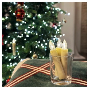 Three warm yellow LED flameless candles in a glass cup with festive Christmas tree background